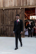Load image into Gallery viewer, Man in a black suit standing in front of a wooden barn door with indoor wedding decorations.