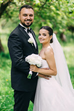 Load image into Gallery viewer, Man in a tuxedo and woman in a wedding dress standing together outdoors with greenery in the background.